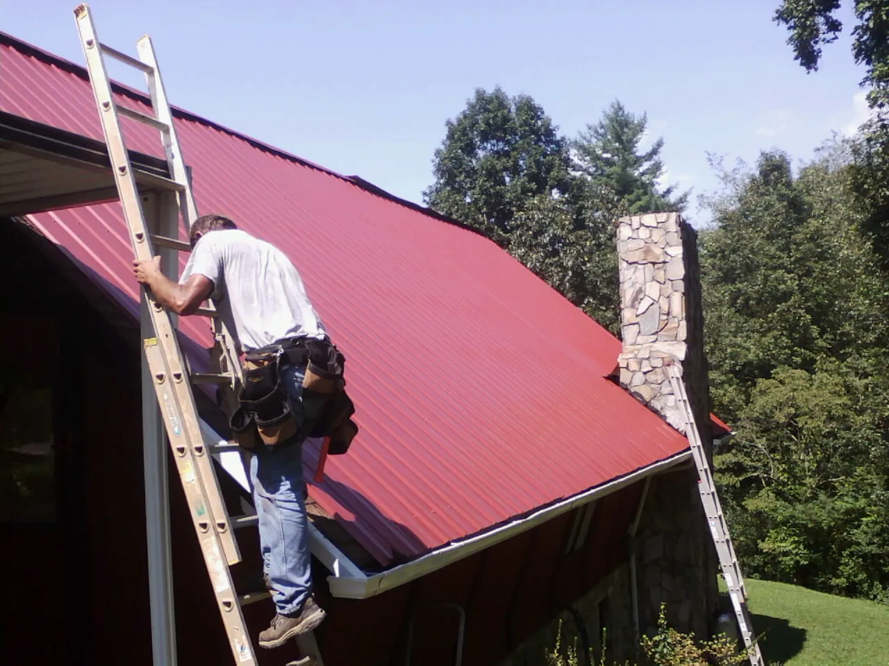 Roofer on a ladder beside a red metal roof and stone chimney — Soffit & Fascia Repair by Roofing Brothers in Westdale